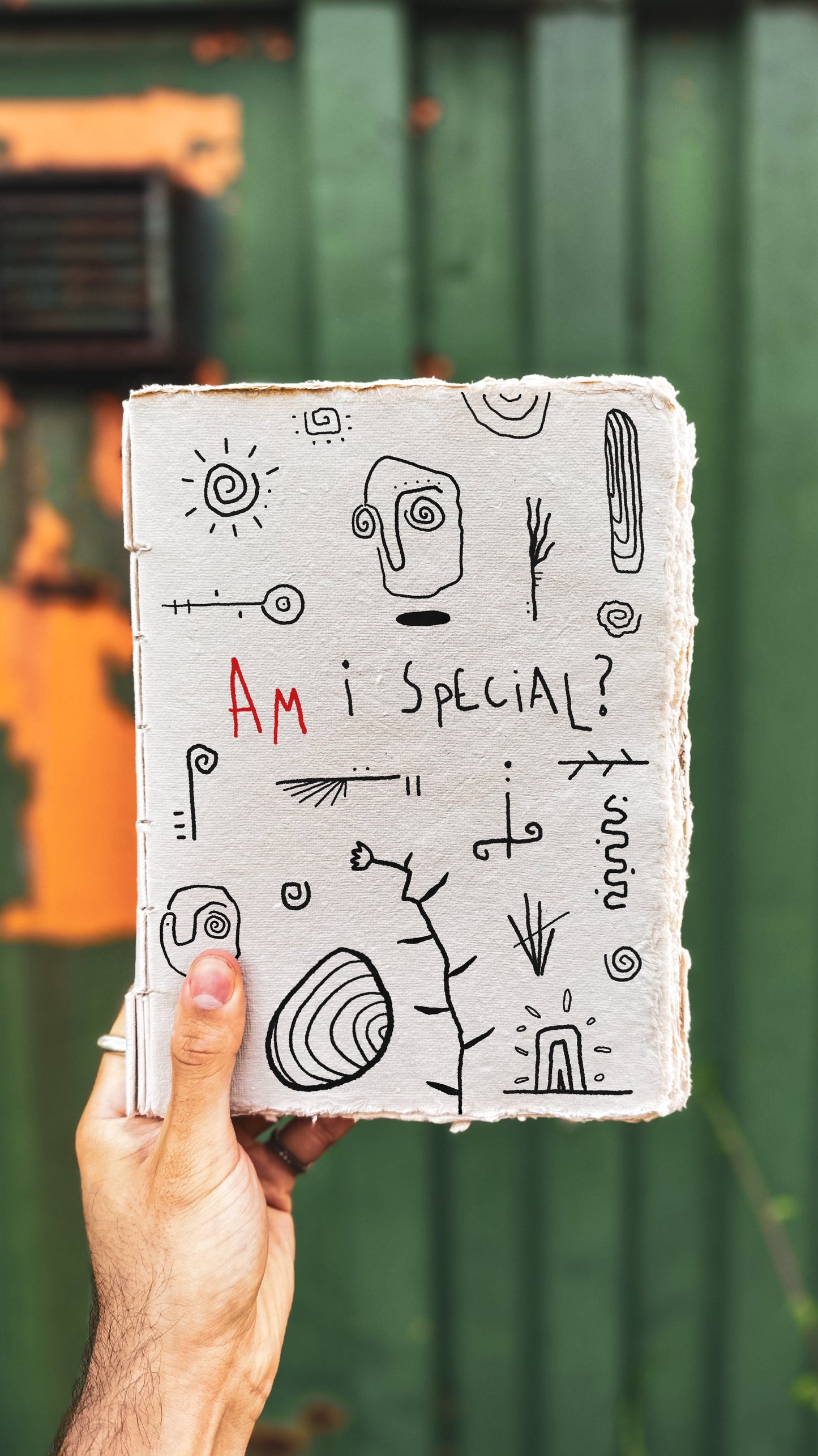 Hand holding a notebook with abstract doodles of symbols and faces and text 'AM I SPECIAL?' against a green wall.