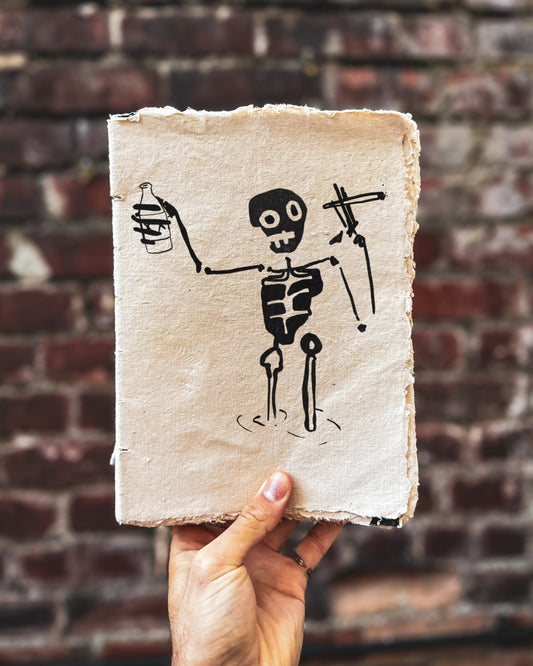 Hand holding a piece of paper with a bold skeleton drawing against a brick wall background. The skeleton is standing in water and holding a bottle and cigarette.